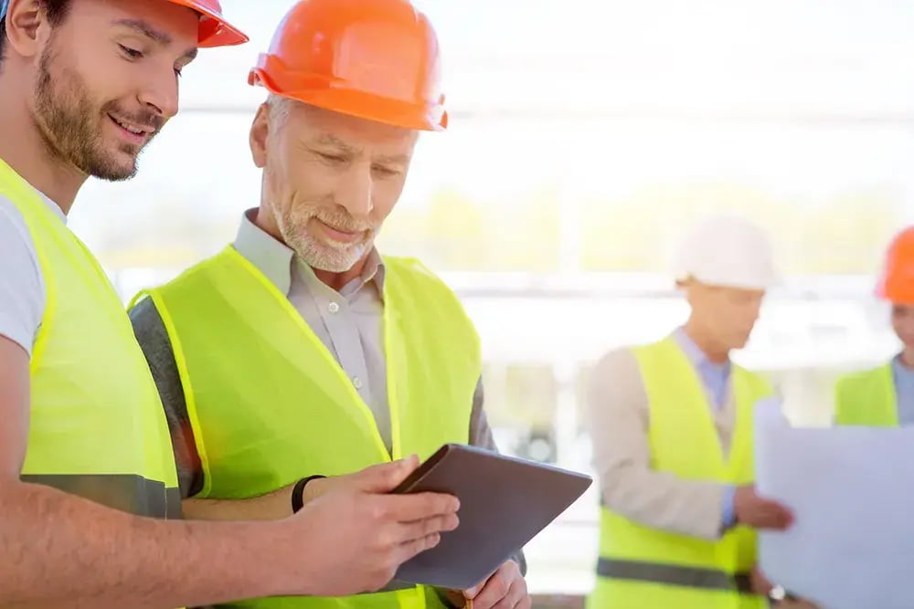 Construction workers in safety vests and hard hats reviewing information on a tablet, with colleagues examining blueprints in the background.