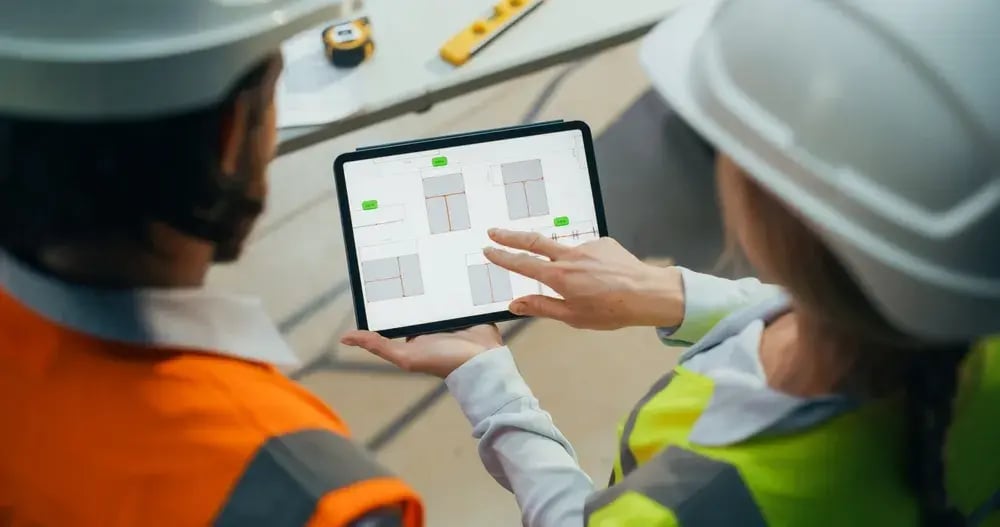 Two construction professionals in hard hats and high-visibility vests review digital building plans on a tablet at a job site.