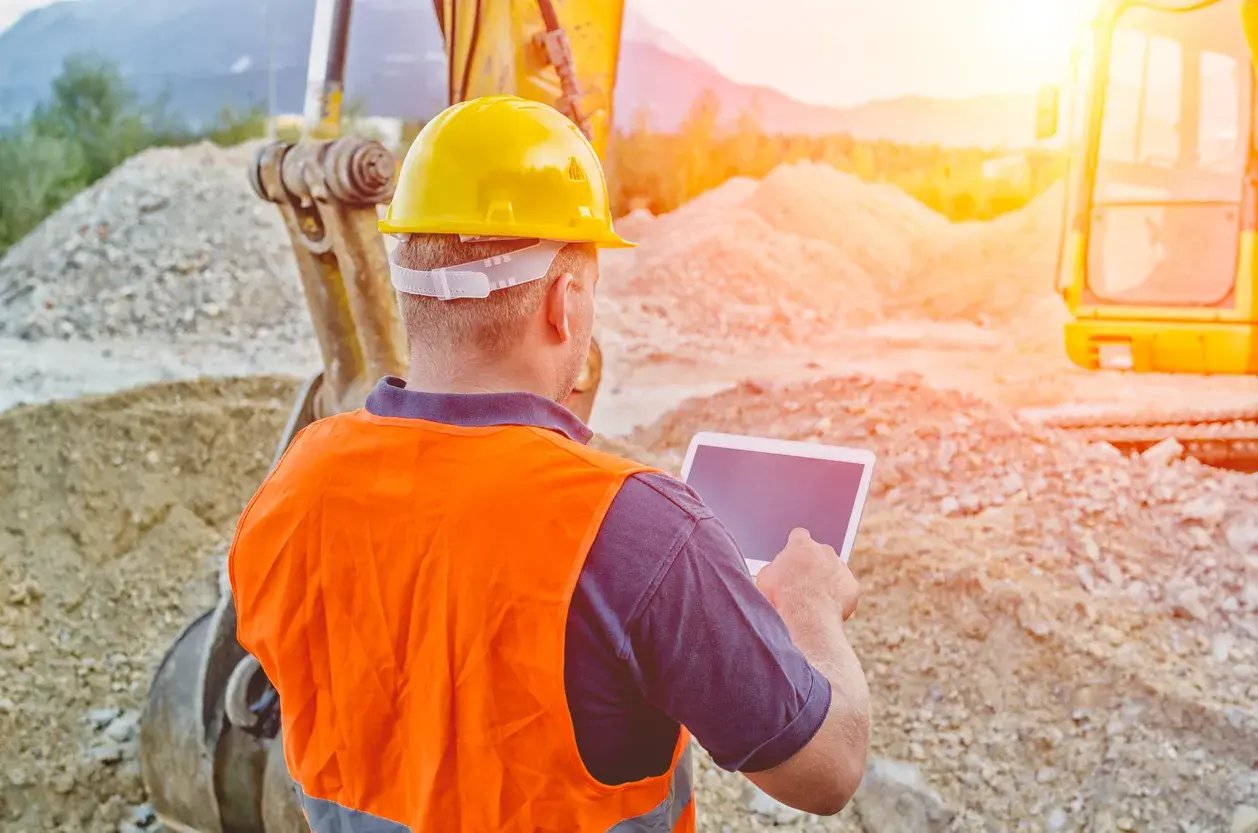 Construction worker in a hard hat and orange safety vest using a tablet beside an excavator at a sunlit construction site.