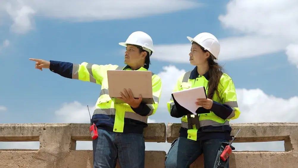 Two construction workers wearing hard hats and safety vests, engaged in a discussion at a construction site.