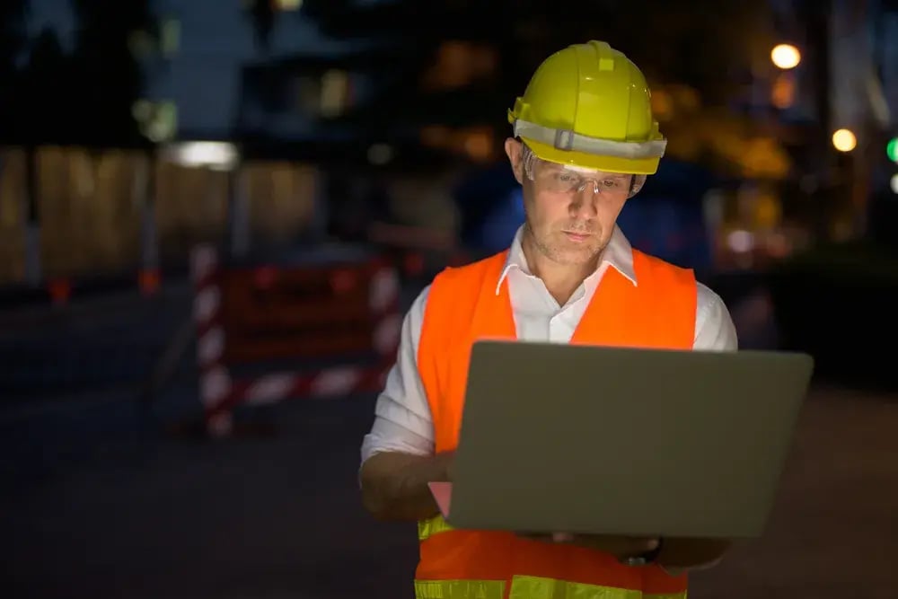 A man wearing a hard hat and safety vest is focused on using a laptop at a construction site.